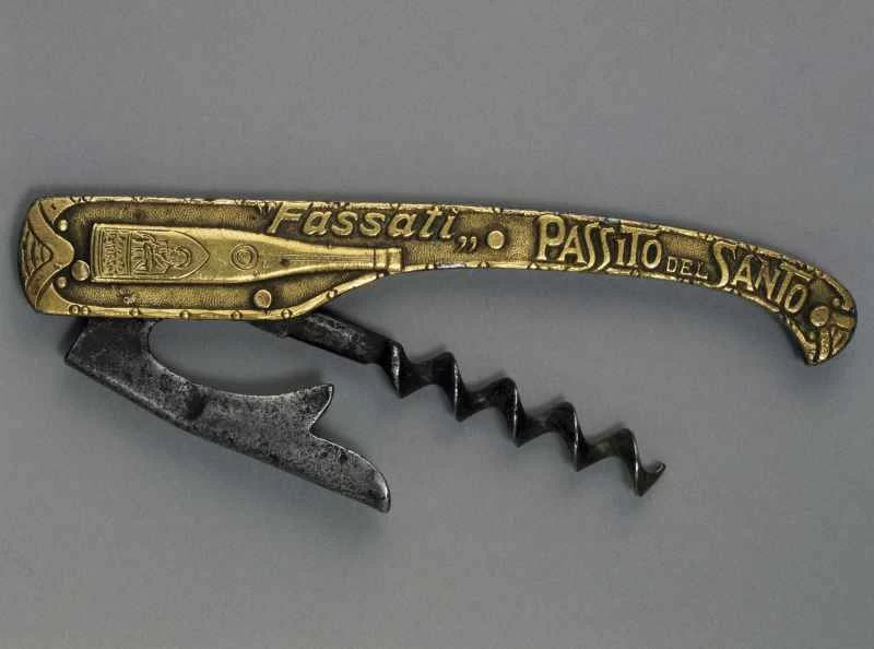 圖中展示的是復古折疊侍者開瓶器(卡爾維恩克設計),背景為中性灰色。手柄採用壓花金色金屬製成,飾有「Fassati Passito del Santo」字樣和酒瓶圖案。手柄展開後,可見深色金屬螺旋鑽頭和帶有凹槽的銀色槓桿臂,展現了早期便攜式開瓶器的機械設計。